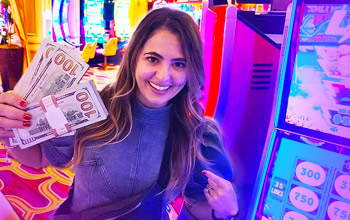 Photograph of Francine Marin aka Lady Luck HQ holding stacks of dollar bills and standing besides a slot machine. 